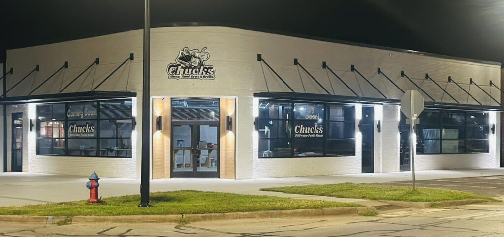 A white brick restaurant called Chucks Public House, lit up at night, sits on a corner with large windows and black awnings. A fire hydrant and street sign are visible near the building, adding to the charm of this local Neighbor-in-the-Know favorite. -WorkIT Coworking Center